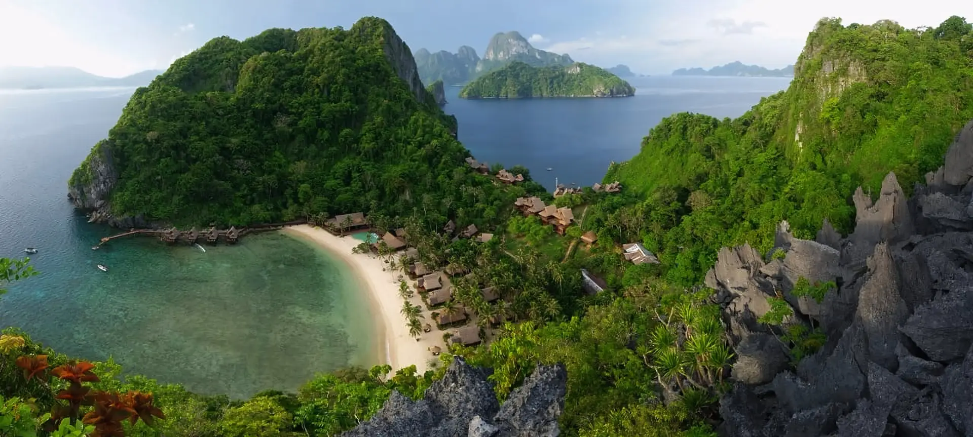 Panoramic view of Cauayan Boutique Private Island from the summit viewdeck, showing the full resort, white sand beach, water villas, reef and Bacuit Bay's limestone islands beyond