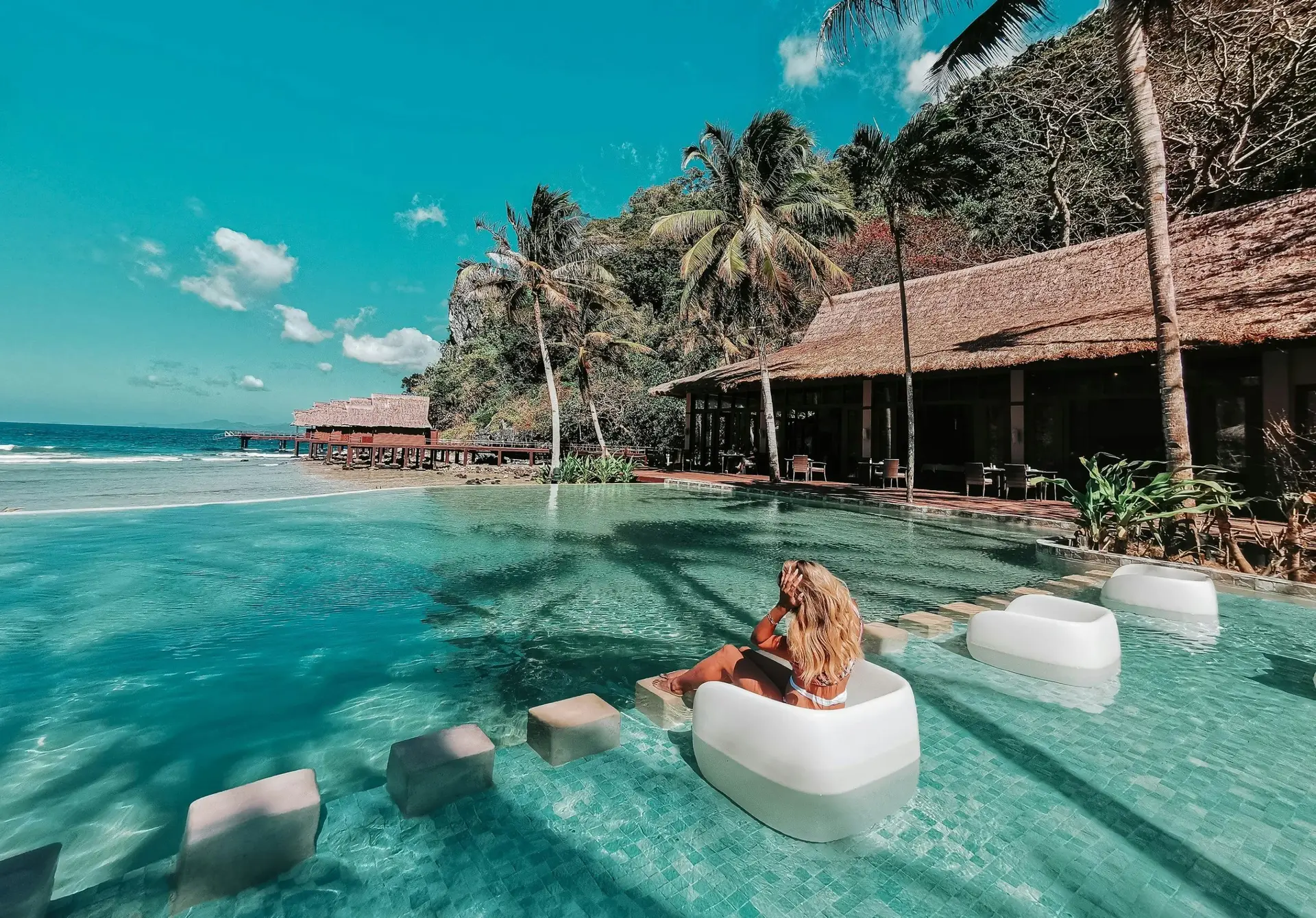 A guest relaxes on a submerged pool lounger in the infinity pool at Cauayan Boutique Private Island, with the beach, water villas and palm trees beyond