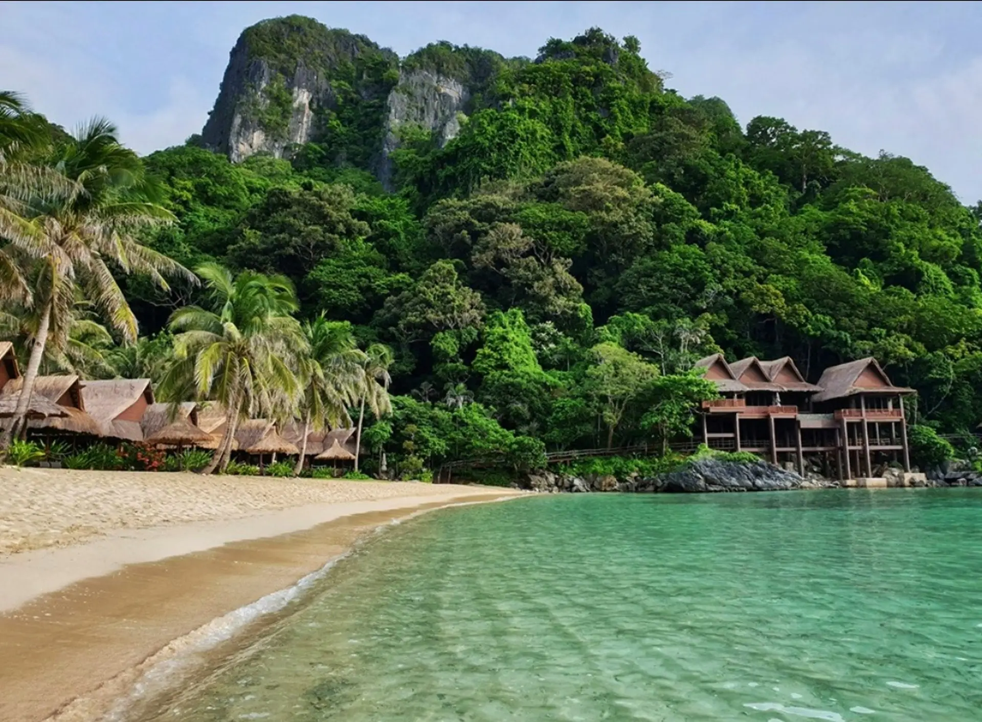 Cauayan Boutique Private Island seen from the water, with thatched beachfront villas along the sand and larger pool villas stepping up the jungle hillside beneath a limestone karst