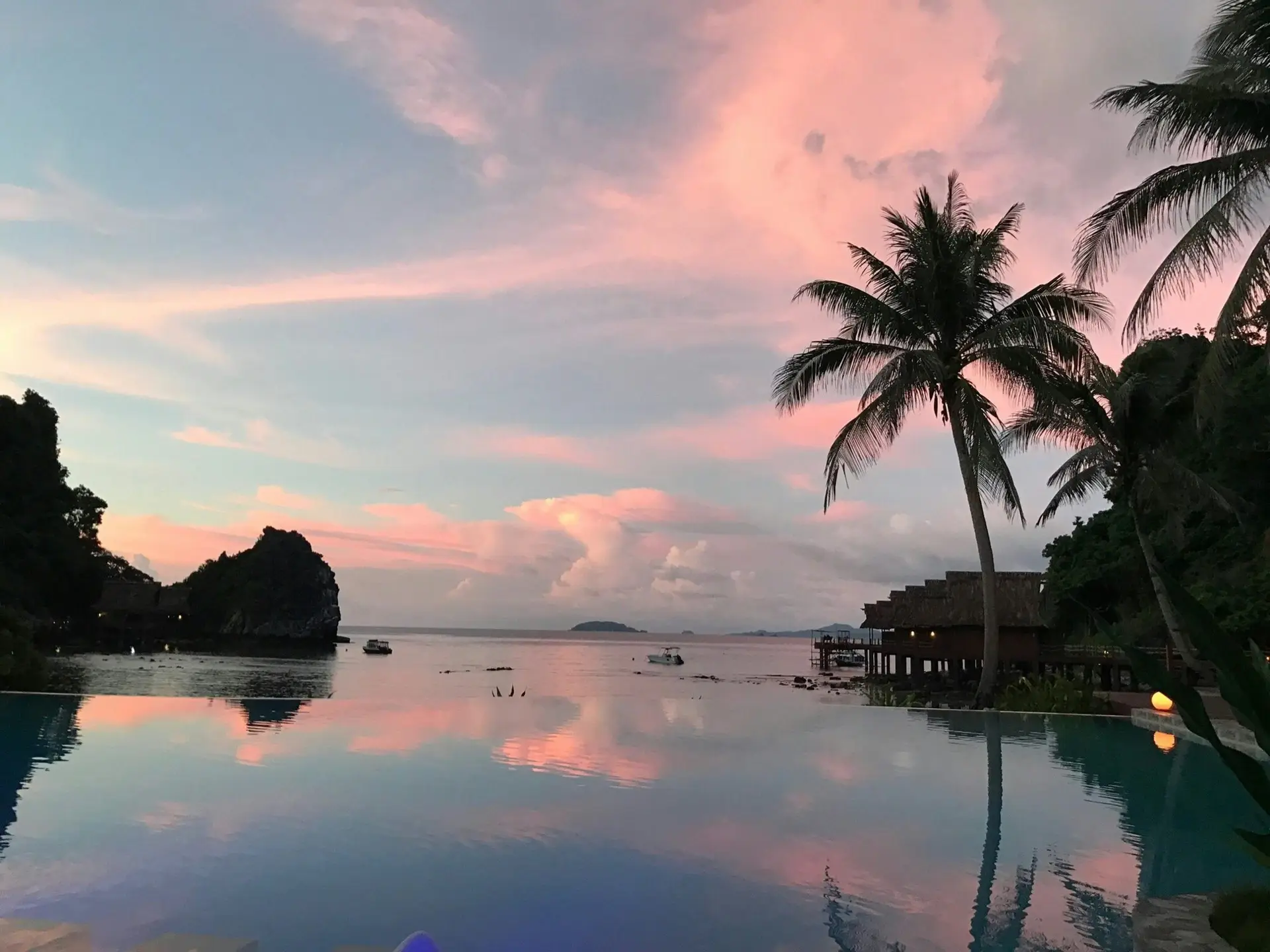 Pink sunset reflected in the infinity pool at Cauayan Boutique Private Island, with a palm tree silhouette, water villas and limestone outcrop framing Bacuit Bay