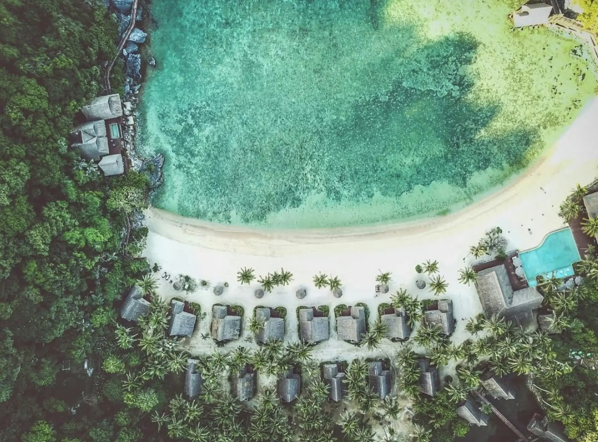 Aerial drone view of Cauayan Boutique Private Island showing beachfront villas along a curved white sand beach, the infinity pool, and water villas on the rocky headland