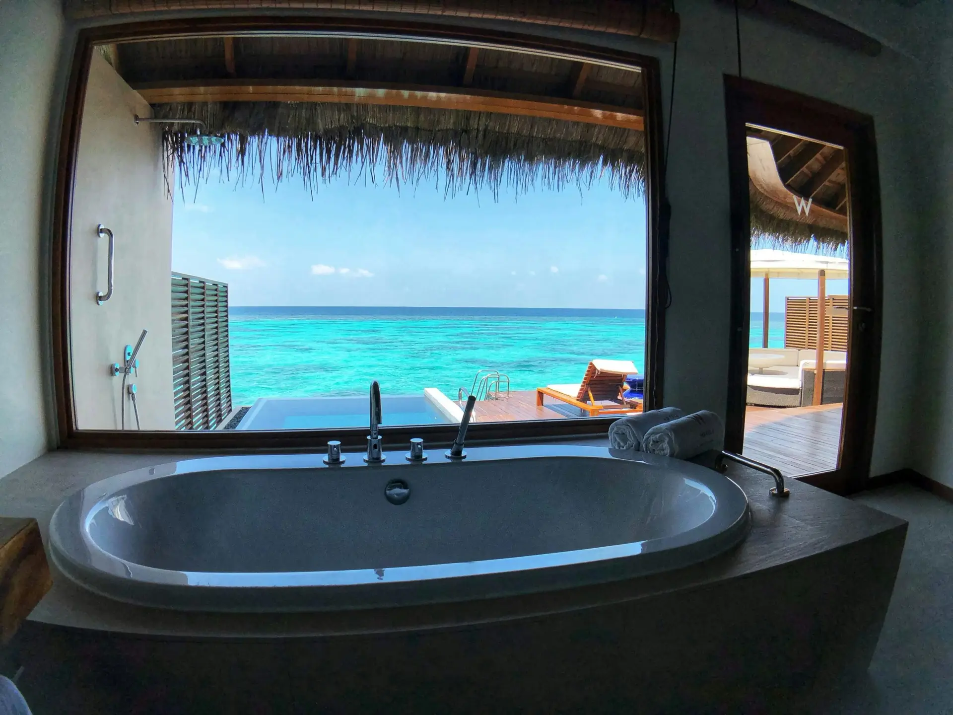 Freestanding bathtub inside an overwater villa at W Maldives looking out through a panoramic window to a turquoise lagoon, private deck with sun loungers and plunge pool