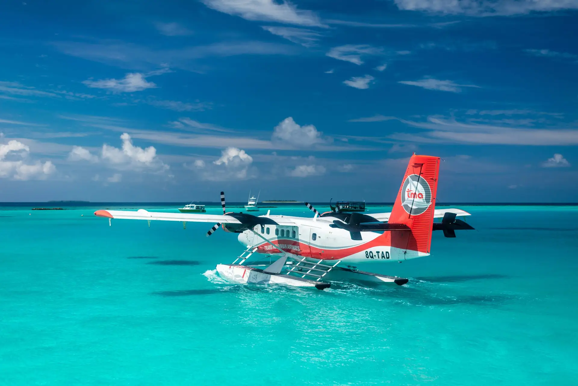 Trans Maldivian Airways Twin Otter seaplane (8Q-TAD) taxiing on turquoise lagoon water with a resort dhoni boat visible in the background