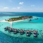Aerial view of a Maldives resort island showing overwater villas on a wooden jetty surrounded by turquoise lagoon water, with beach villas and palm trees on the island behind