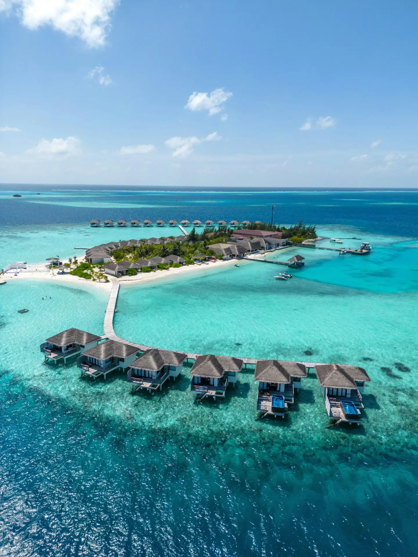 Aerial view of a Maldives resort island showing overwater villas on a wooden jetty surrounded by turquoise lagoon water, with beach villas and palm trees on the island behind