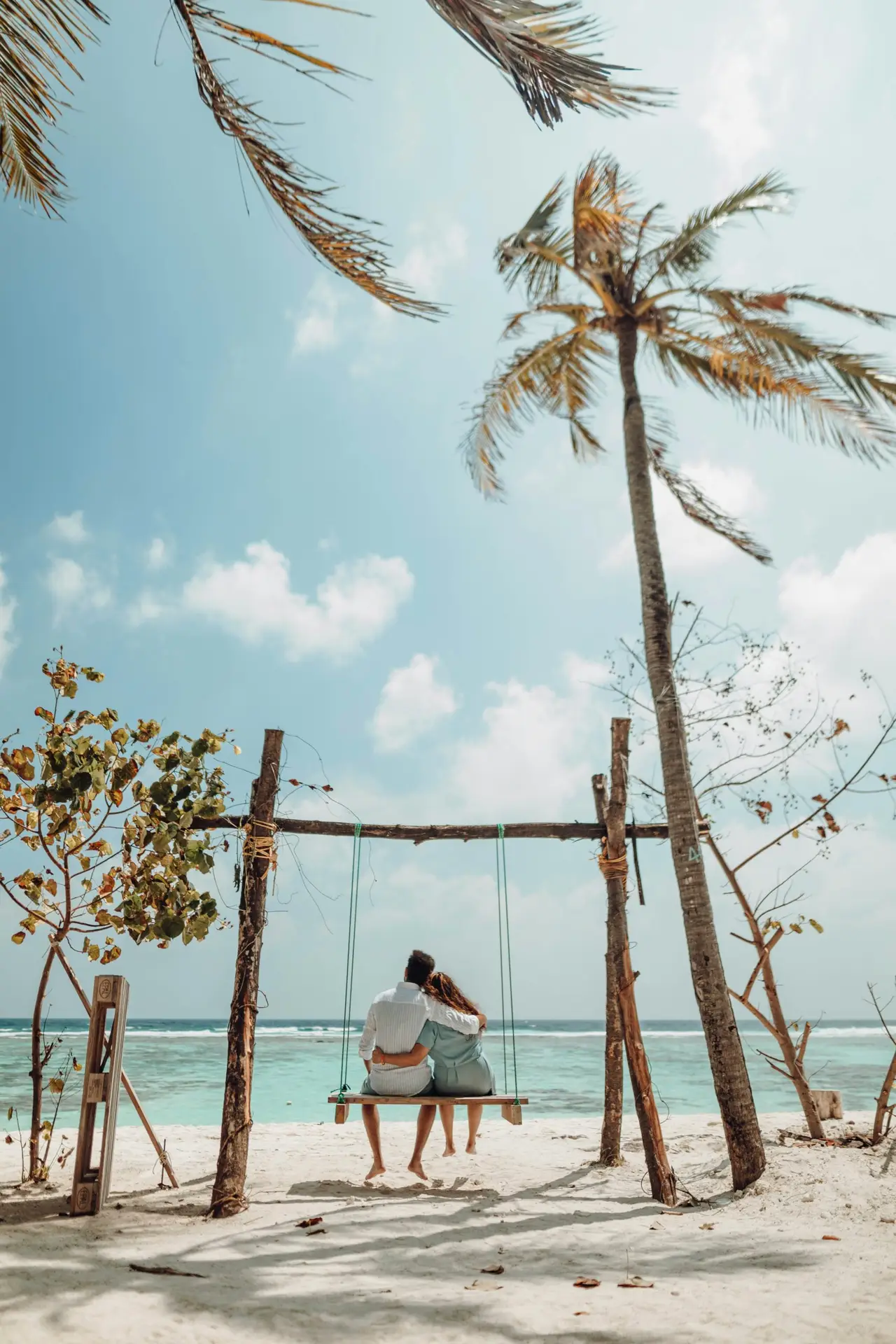 Couple sitting together on a wooden beach swing facing the turquoise ocean under a coconut palm in the Maldives