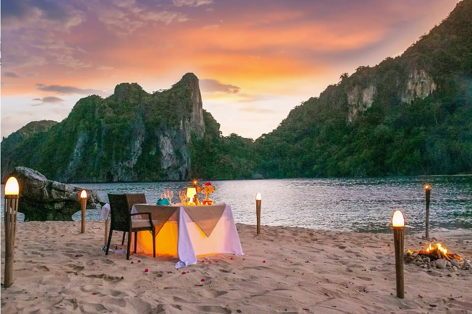 Private beach dinner for two at sunset on Cauayan Island, with tiki torches, a small bonfire, rose petals in the sand and Bacuit Bay karsts glowing orange behind