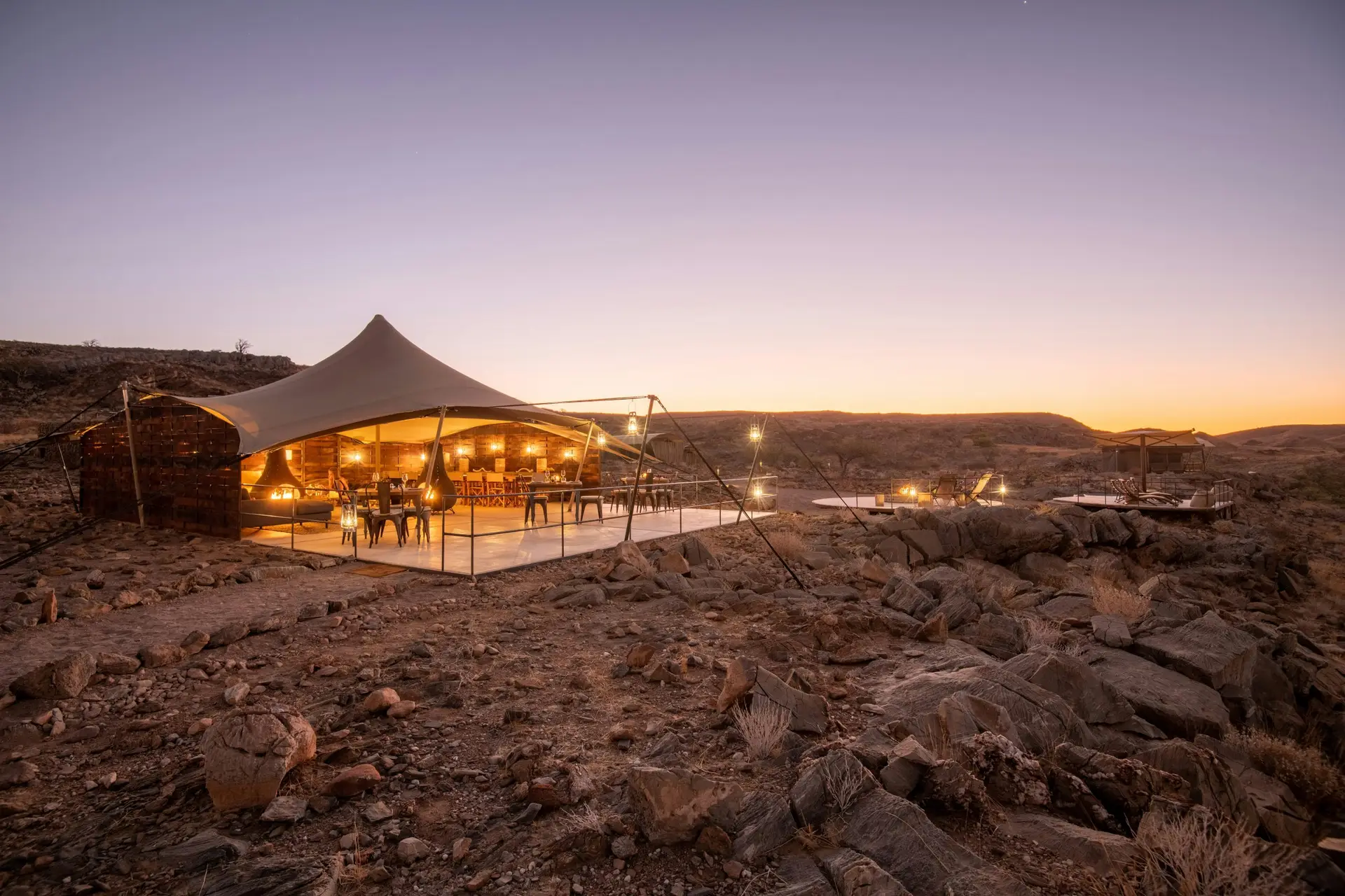 Alt Text: Camp Doros common area and dining tent at dusk, perched on a rocky ridge above a dry riverbed in Damaraland, Namibia
