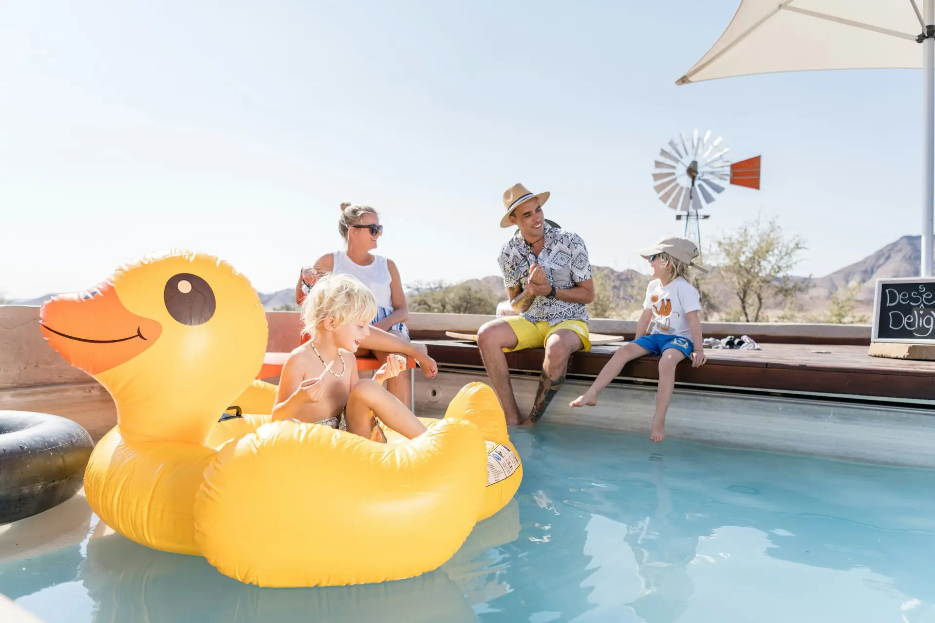 A family relaxing by the pool at Camp Sossus, with a child in an inflatable duck, a windmill and Namibian mountains in the background