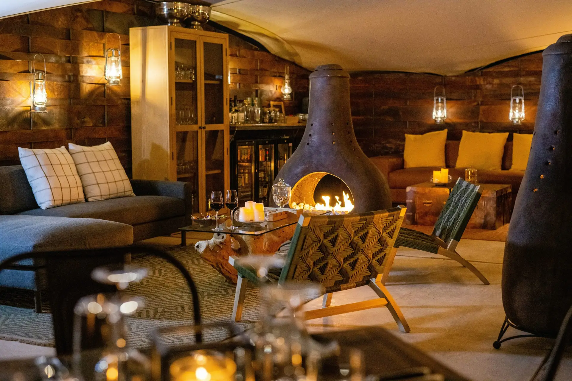 The lounge at Onduli Ridge in the evening, with a chiminea fireplace, lanterns, wine glasses and woven chairs against granite walls