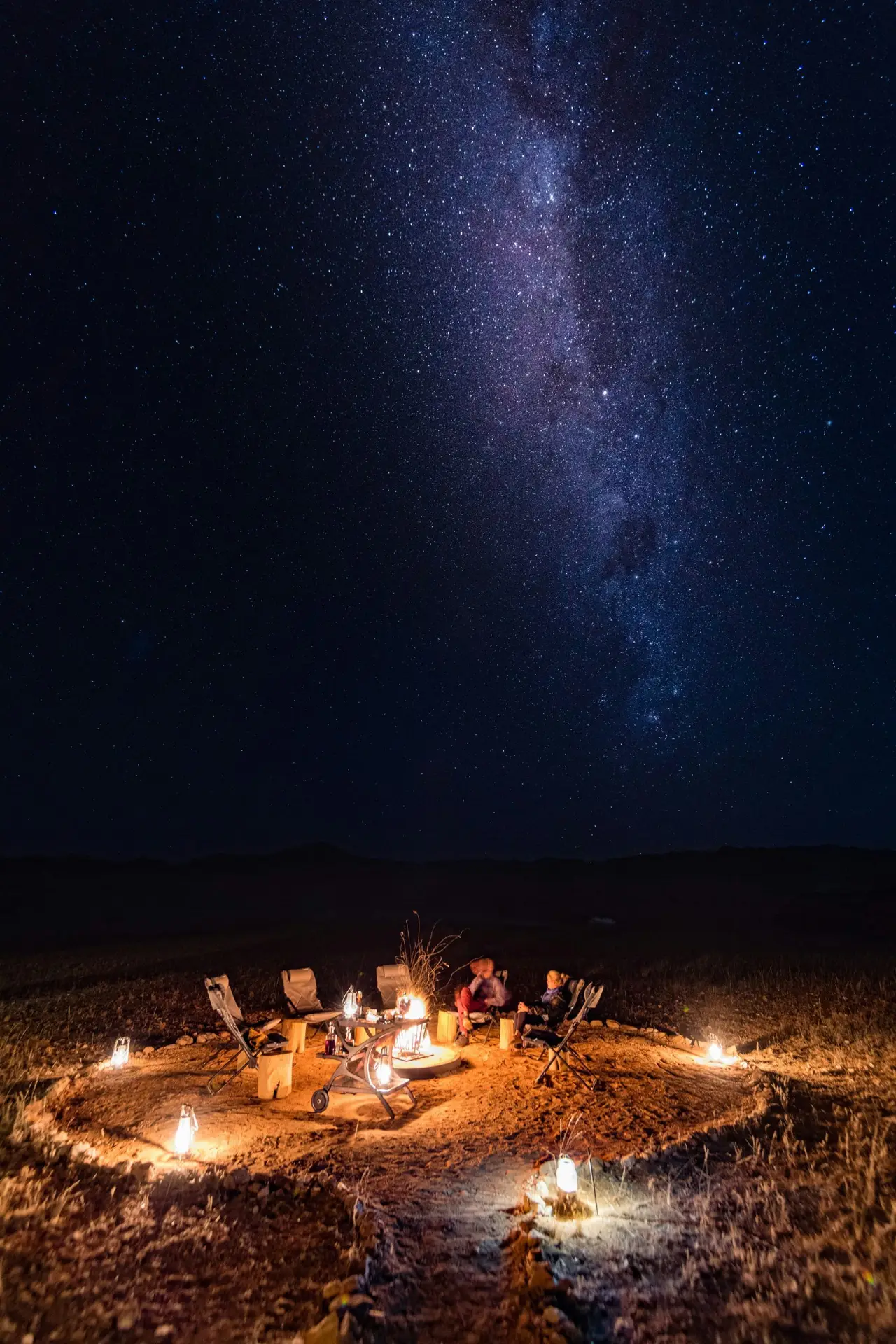 Guests seated around a campfire under the Milky Way at Onduli Ridge, Damaraland, with lanterns lighting the desert floor