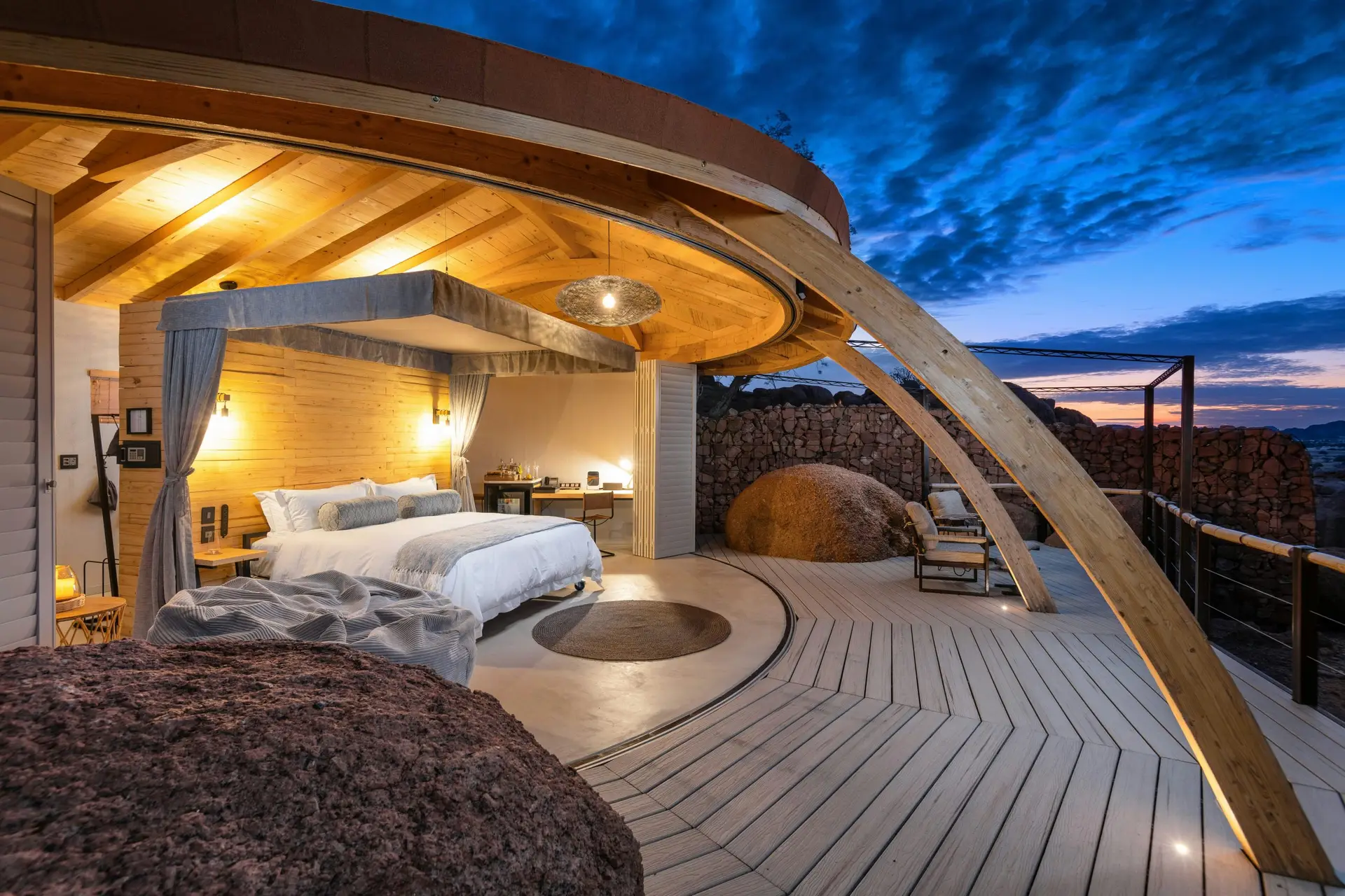  Suite interior at Onduli Ridge at dusk, with a king-size bed on rails opening onto a wooden deck built around granite boulders