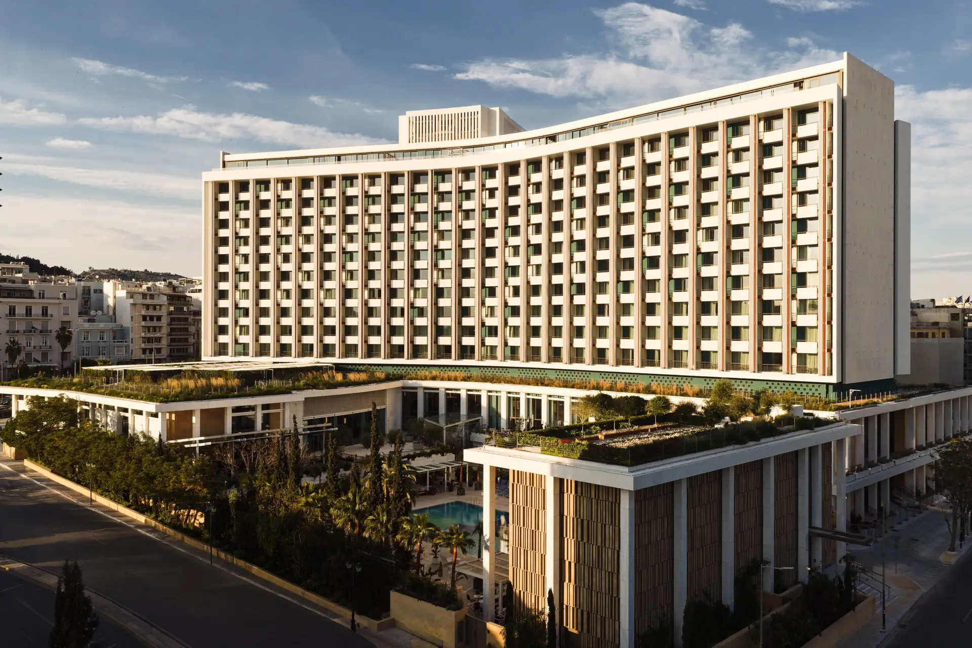 Exterior of Conrad Athens The Ilisian in Ilisia, showing the curved modernist facade of the former Hilton Athens building above the pool courtyard and green roof gardens 