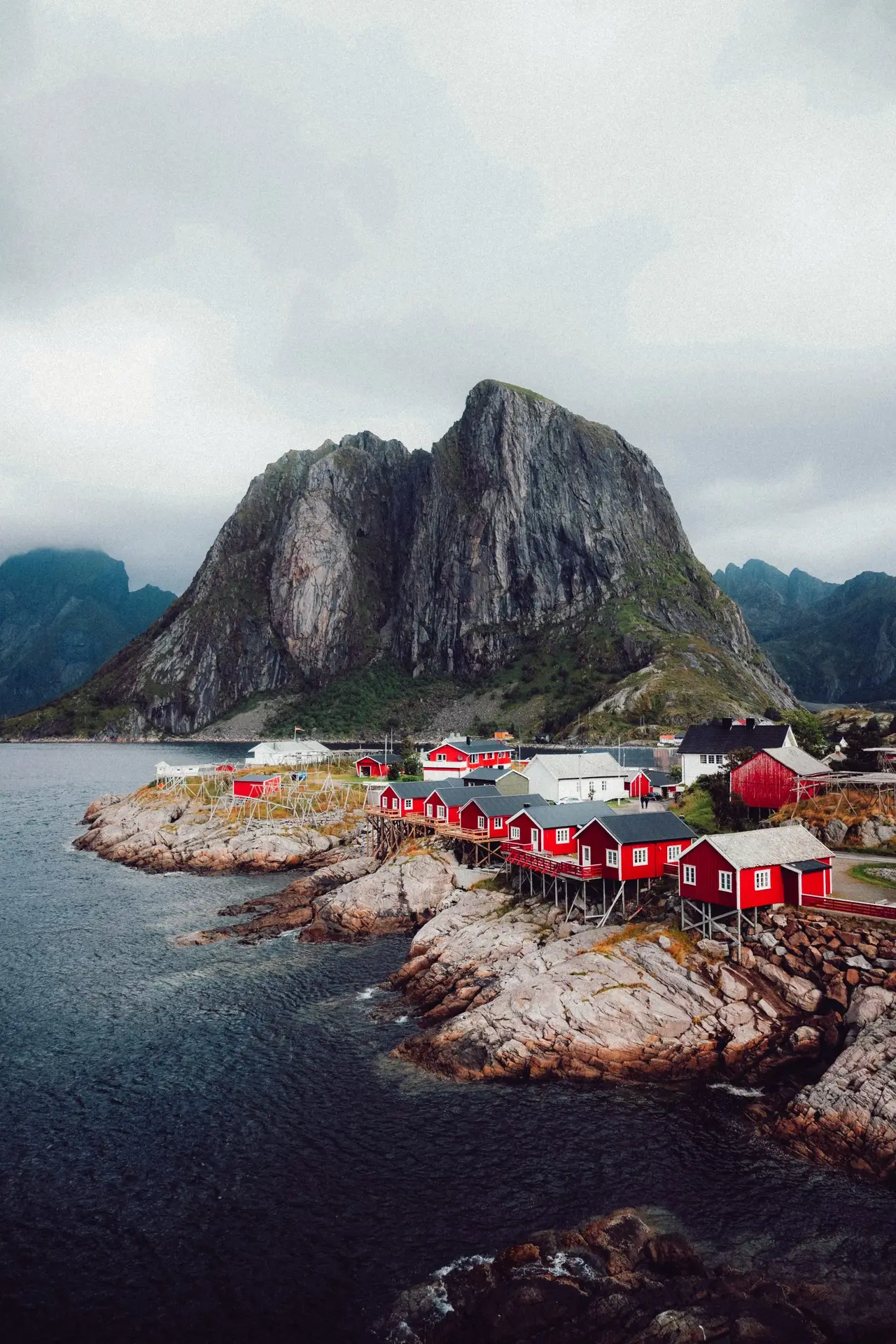 Traditional red rorbuer fishing cabins on stilts cluster on the rocky shoreline of Hamnøy in Lofoten, Norway, beneath dramatic granite peaks under a moody sky.