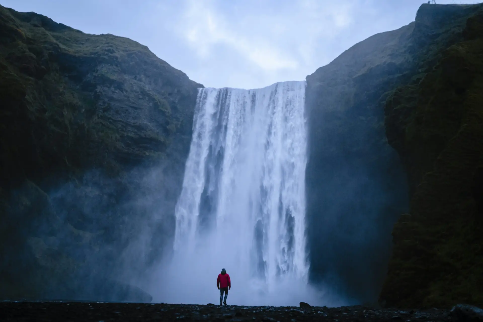Solo travel destinations 2026: a solo traveler in a red jacket stands at the base of Skógafoss waterfall in Iceland, the world's safest country and the second-strongest first-timer pick of the year