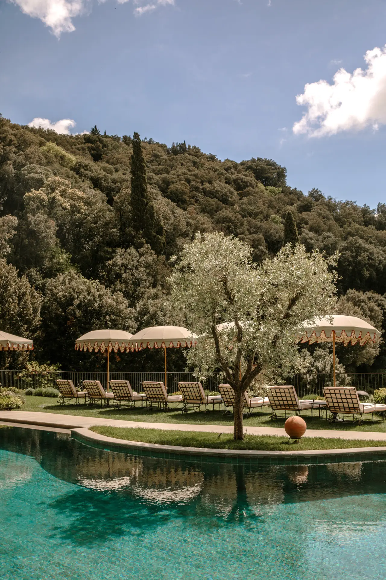 The heated swimming pool at Villa San Michele in Fiesole with olive tree, Belmond-signature umbrellas, and the Leonardo Woodland in the background