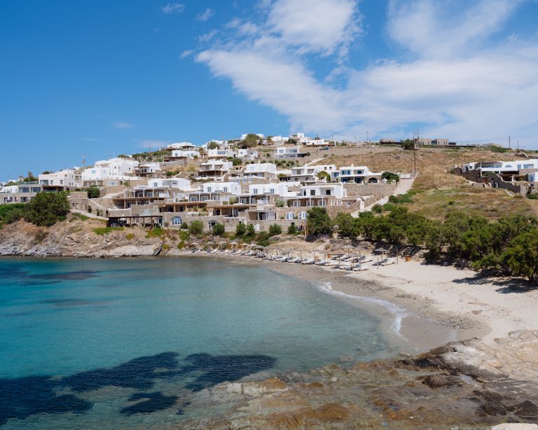 Casa Del Mar Mykonos review: the villas seen from across Glyfadi cove, whitewashed and stepping down the hillside above two rows of thatched umbrellas and sun loungers on the sandy beach