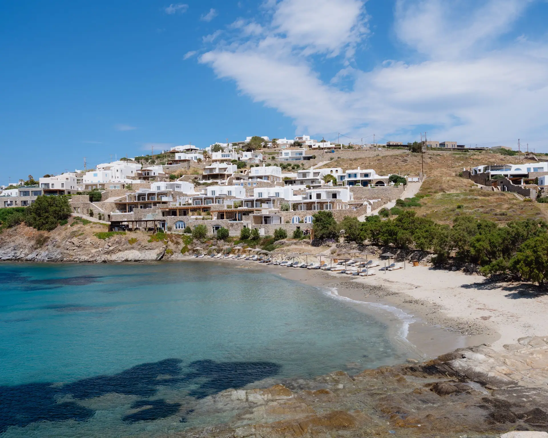Casa Del Mar Mykonos review: the villas seen from across Glyfadi cove, whitewashed and stepping down the hillside above two rows of thatched umbrellas and sun loungers on the sandy beach