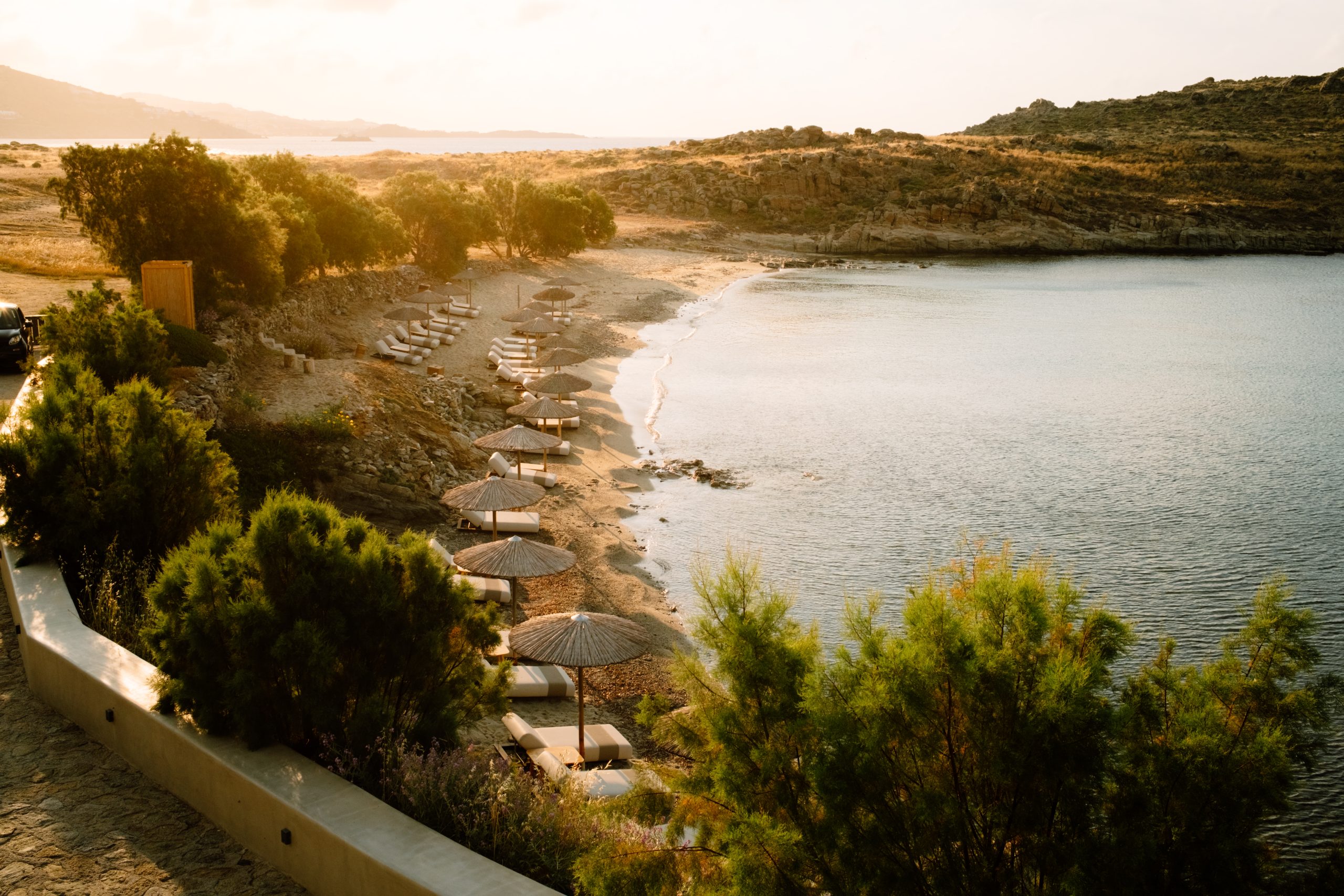 Casa Del Mar Mykonos: A Week on the Quiet Side of the Island 2 Glyfadi cove at Casa Del Mar Mykonos in golden sunrise light, with empty thatched umbrellas and loungers along the sand and the calm, mirror-still sea