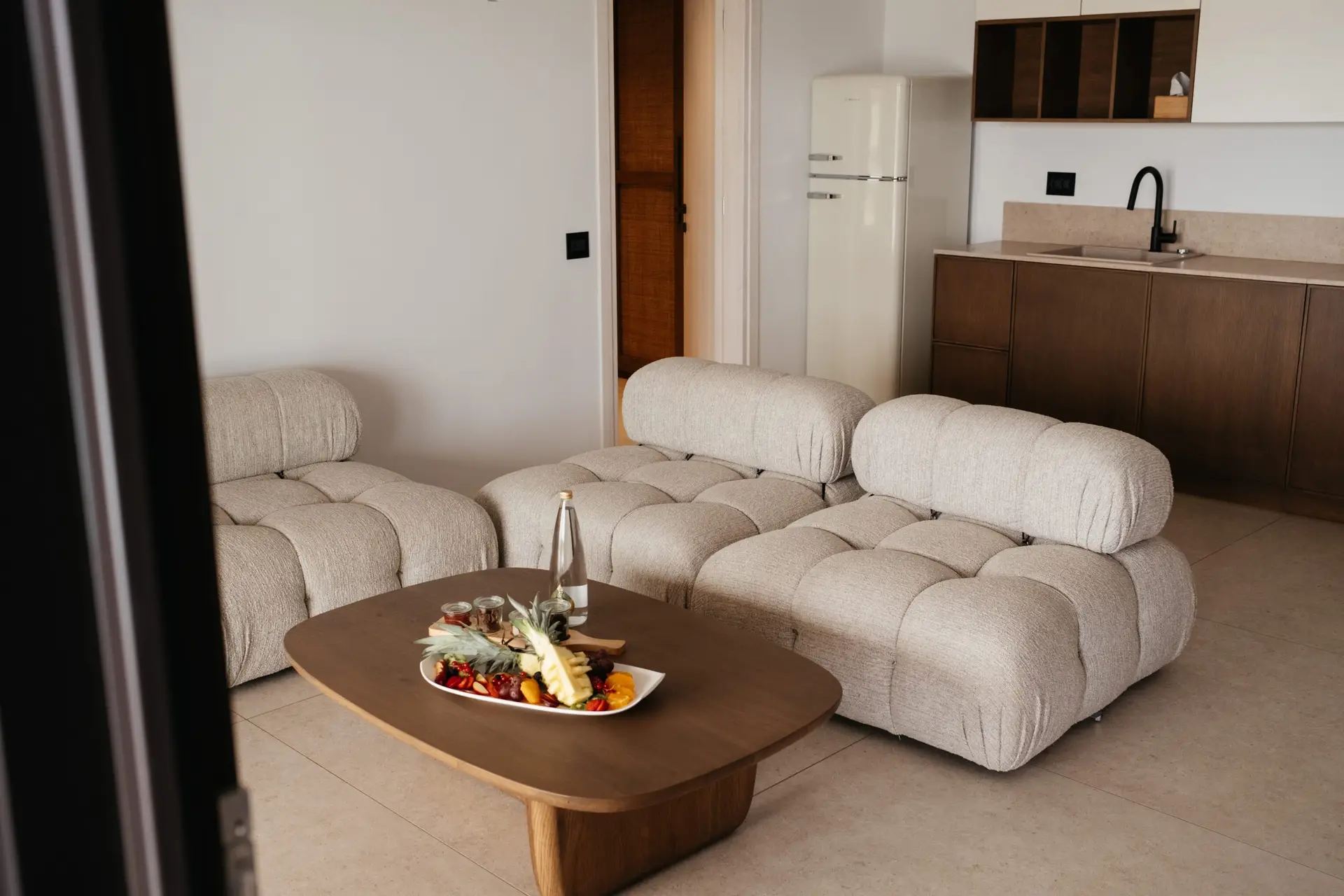 Casa Del Mar Mykonos: A Week on the Quiet Side of the Island 7 Open-plan living room in the Royal Beachfront villa at Casa Del Mar Mykonos, with a designer bouclé modular sofa in the Camaleonda style, dark wood coffee table and kitchen cabinetry, and a welcome fruit platter