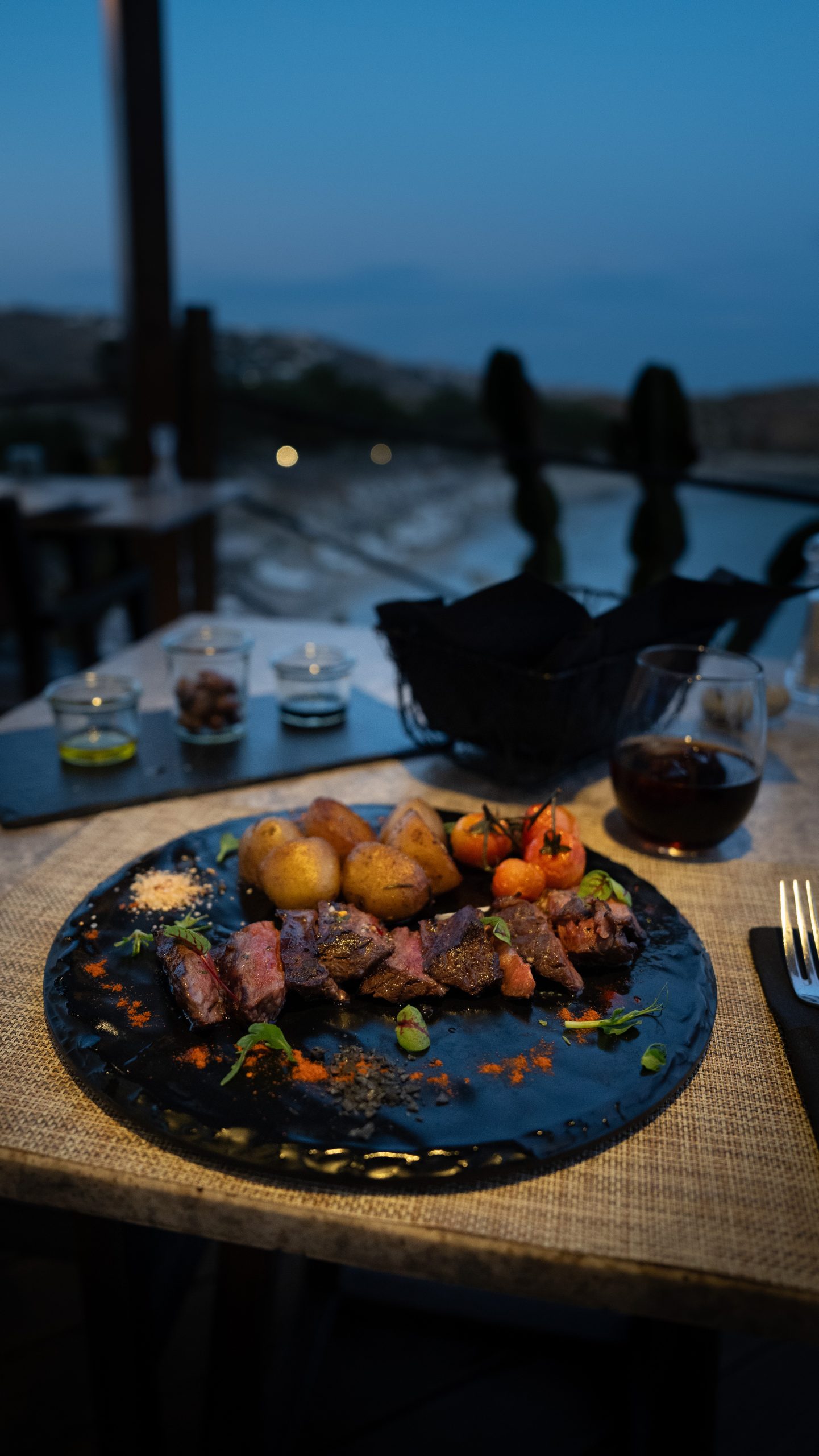 Casa Del Mar Mykonos: A Week on the Quiet Side of the Island 10 Plated beef tagliata with roasted potatoes and cherry tomatoes on a dark slate plate at Miramar restaurant, Casa Del Mar Mykonos, with a glass of red wine and the cove in the blue dusk behind