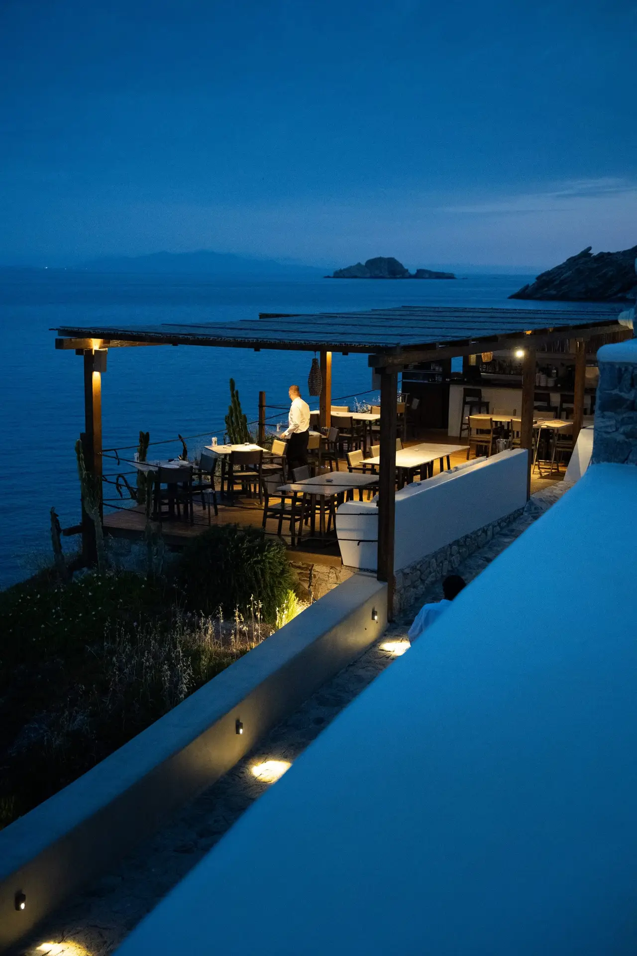 Casa Del Mar Mykonos: A Week on the Quiet Side of the Island 8 The terrace of Miramar restaurant at Casa Del Mar Mykonos at blue-hour dusk, with tables set above the sea under a wooden pergola and a member of staff attending the floor
