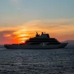 Four Seasons luxury yacht in silhouette on open water at sunset, with the Four Seasons Yacht name on the hull and a dark mountain ridge on the horizon.