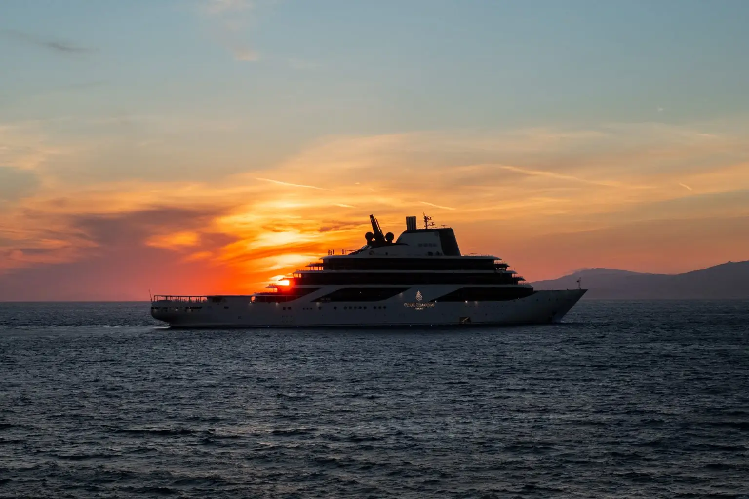 Four Seasons luxury yacht in silhouette on open water at sunset, with the Four Seasons Yacht name on the hull and a dark mountain ridge on the horizon.