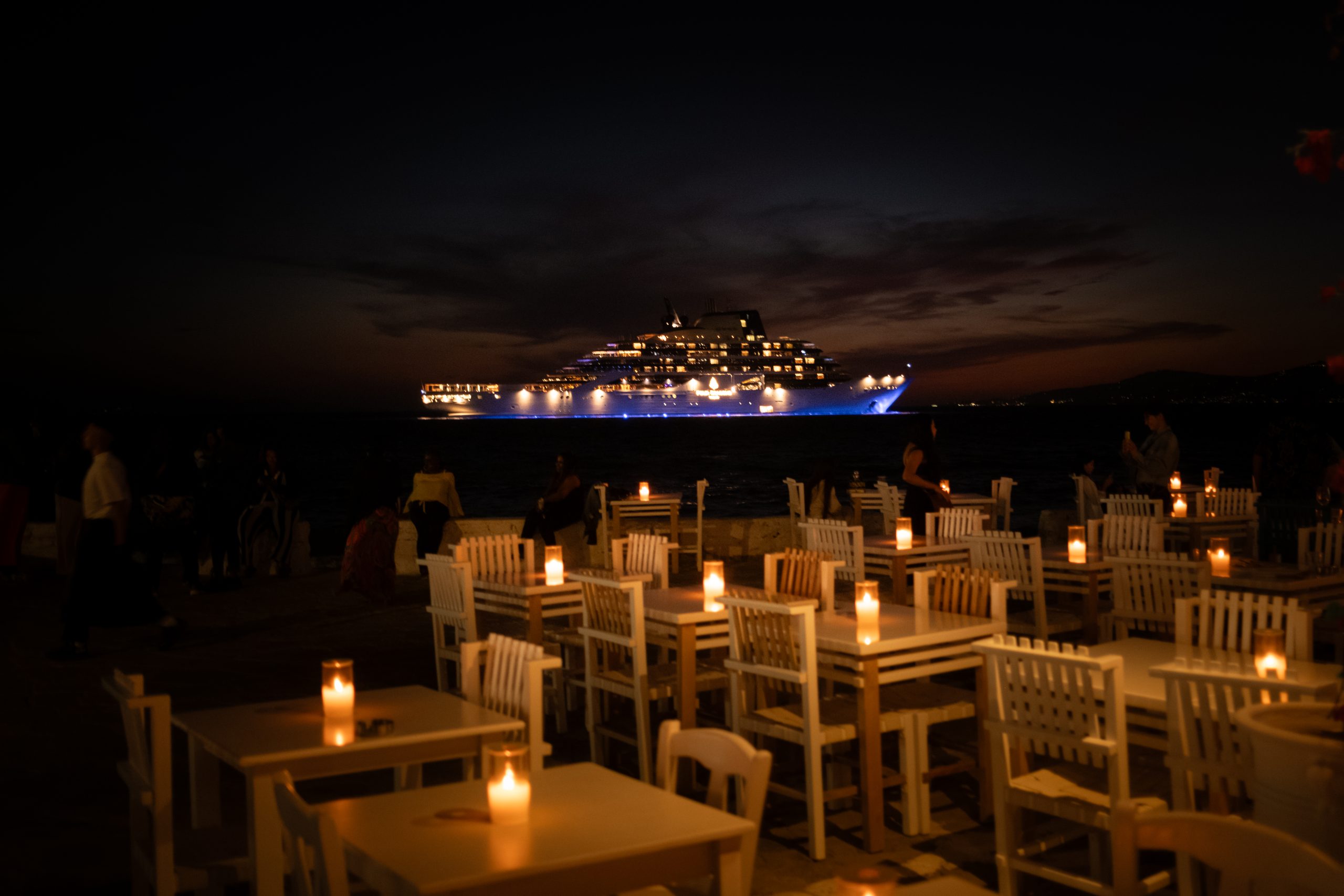 Four Seasons is already building yacht number two. That tells you everything. 4 Four Seasons lit up offshore at night in Mykonos, viewed from a candlelit restaurant terrace with white tables and chairs in the foreground.