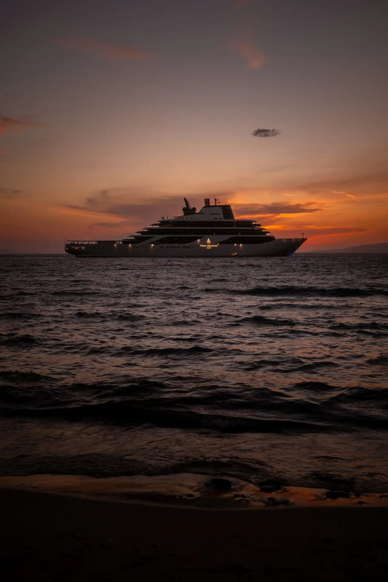 Four Seasons is already building yacht number two. That tells you everything. 1 Four Seasons yacht offshore at sunset in Mykonos, seen from a dark beach with small waves in the foreground.