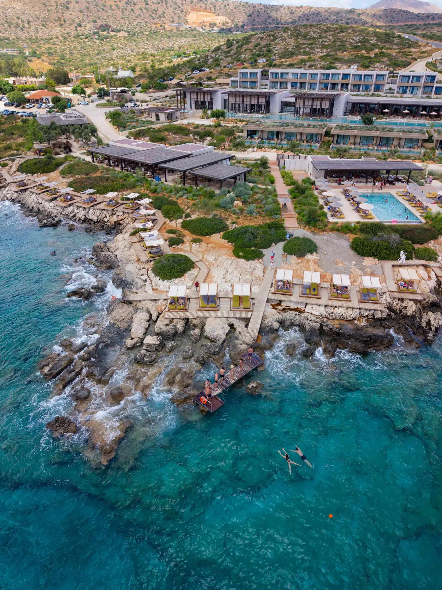 JW Marriott Crete Resort & Spa: The Complete Guide to Greece’s First JW Marriott 7 Aerial view of the JW Marriott Crete seafront, with cabanas along the rocks, guests swimming from the pier, and clear turquoise water