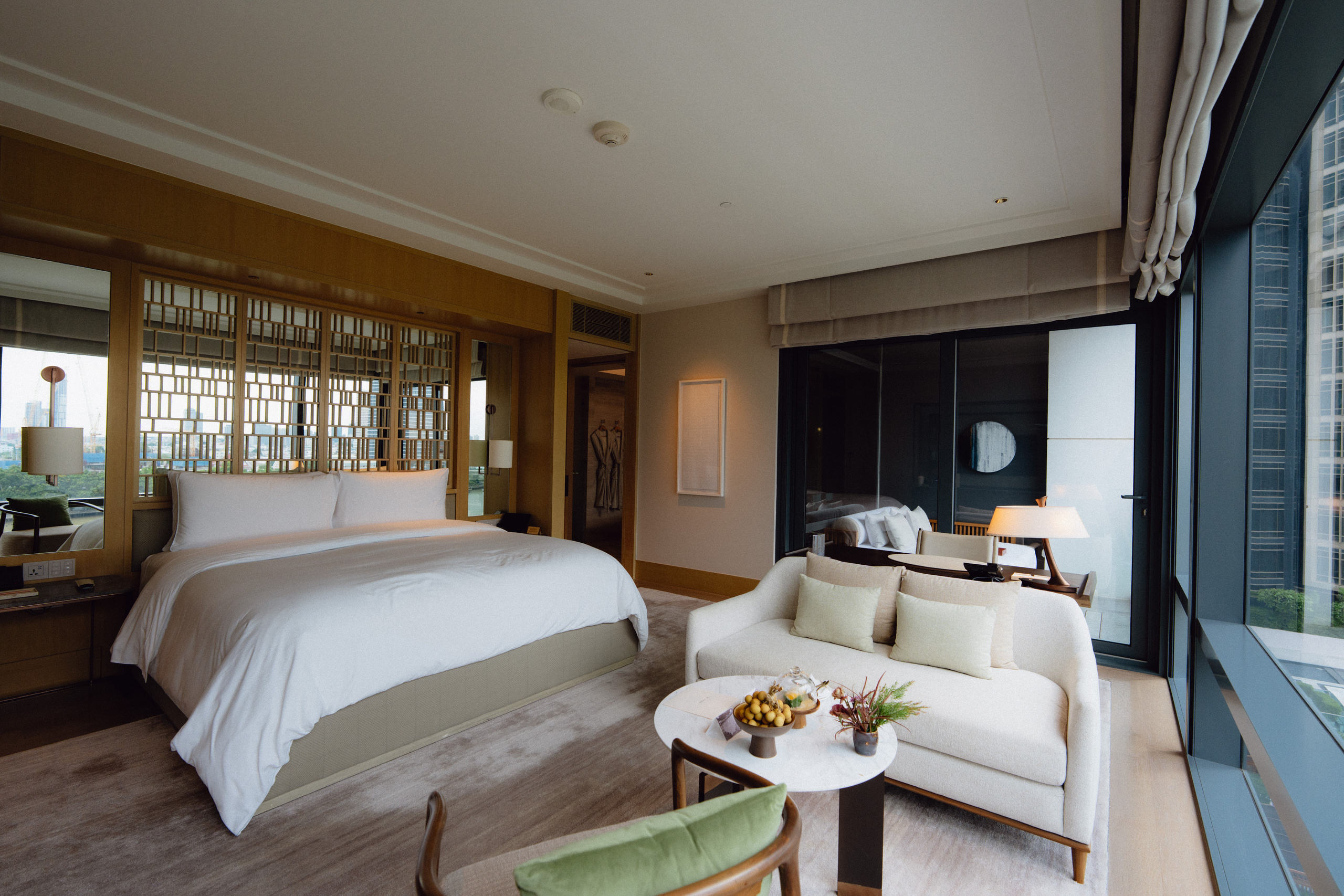 King bed with a wooden lattice headboard and a sofa by the window in a Riverfront Premier room at Capella Bangkok