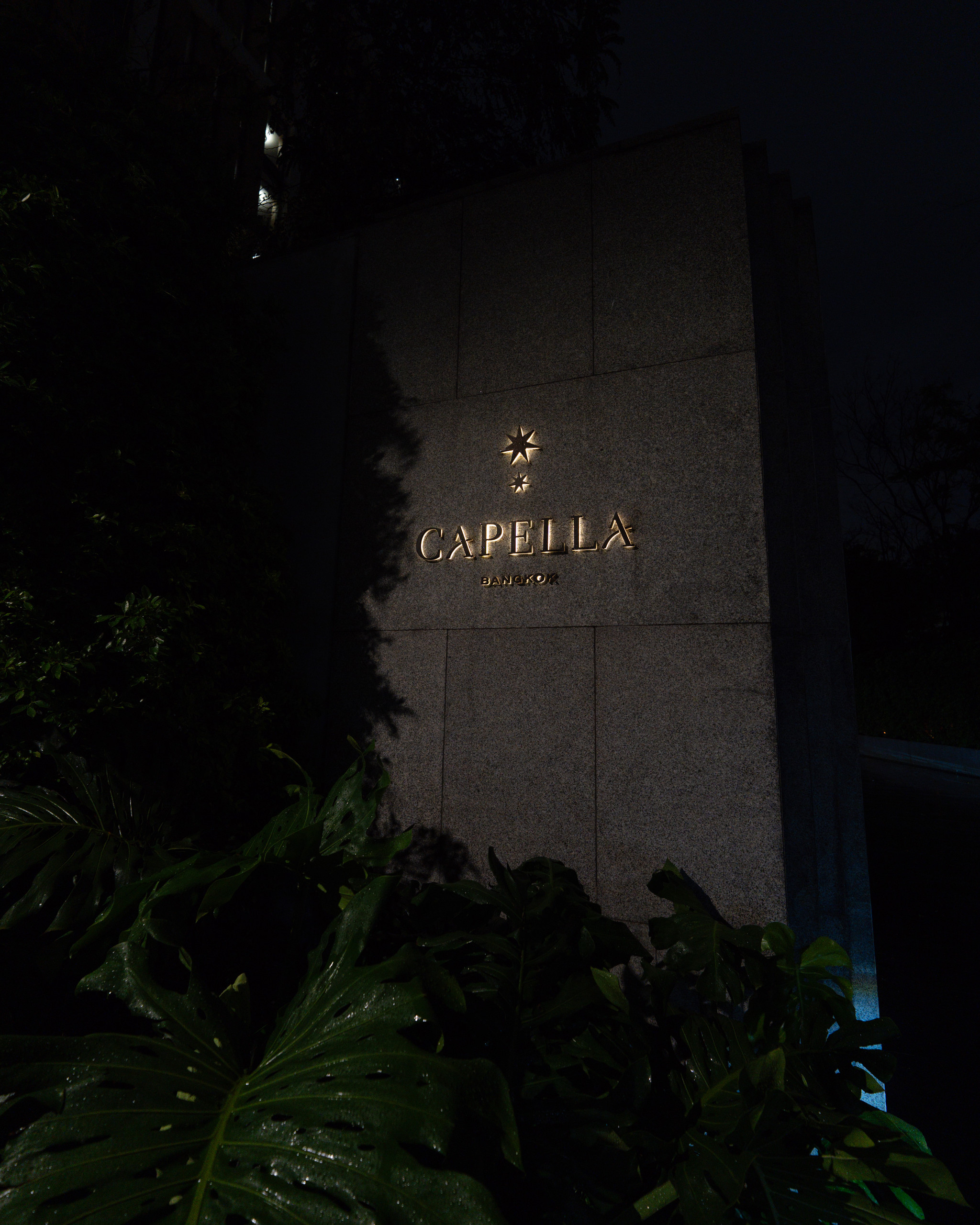 The illuminated Capella Bangkok sign at night, framed by tropical leaves