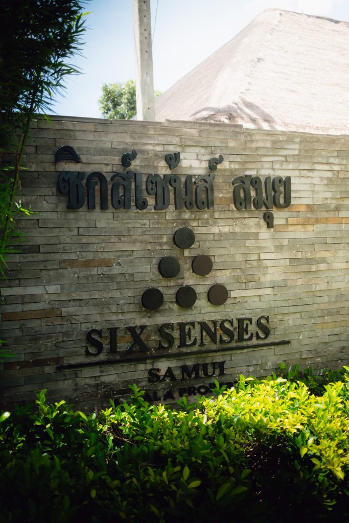 Stone Six Senses Samui entrance sign with Thai script and the logo, framed by green plants